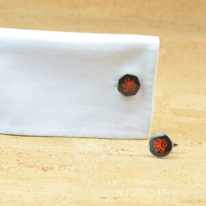 May include: A pair of cufflinks with a red lion design on a black background. The cufflinks are on a white shirt cuff and a cork surface.
