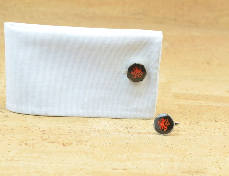 May include: A pair of cufflinks with a red lion design on a black background. The cufflinks are on a white shirt cuff and a cork surface.