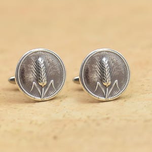 May include: Silver cufflinks featuring a wheat stalk design. Each cufflink has a circular face with a detailed relief of a wheat stalk. The cufflinks have a classic, elegant appearance. The background is textured.