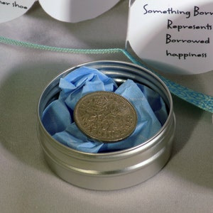 Puede incluir: Una peque&ntilde;a lata de plata con un pa&ntilde;uelo azul en su interior, que contiene una moneda de plata. La lata est&aacute; etiquetada como "Something Borrowed" y dice "Represents Borrowed happiness".