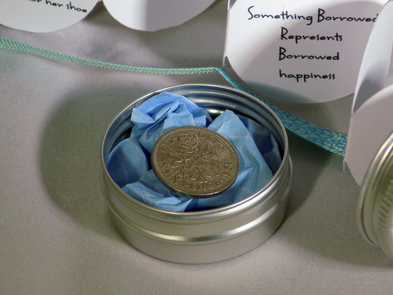 Puede incluir: Una peque&ntilde;a lata de plata con un pa&ntilde;uelo azul en su interior, que contiene una moneda de plata. La lata est&aacute; etiquetada como "Something Borrowed" y dice "Represents Borrowed happiness".