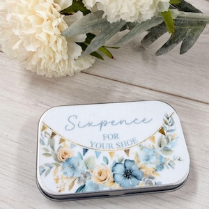 May include: A rectangular tin with a floral design and the text "Sixpence FOR YOUR SHOE". The design features blue and peach flowers with green leaves and a gold banner. The tin has a silver edge.