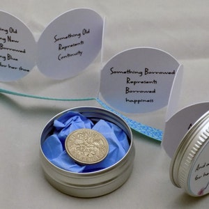 Puede incluir: Una peque&ntilde;a lata de plata con una tapa con estampado azul y blanco. La tapa tiene el texto "Something Old, Something New, Something Borrowed, Something Blue & a sixpence for her shoe" impreso en ella. Dentro de la lata hay un papel de seda azul y una moneda de plata.