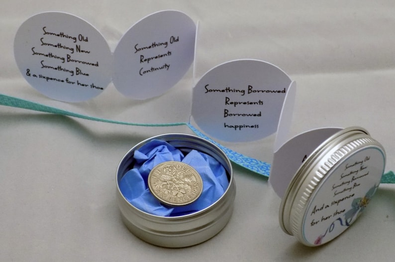 Puede incluir: Una peque&ntilde;a lata de plata con una tapa con estampado azul y blanco. La tapa tiene el texto "Something Old, Something New, Something Borrowed, Something Blue & a sixpence for her shoe" impreso en ella. Dentro de la lata hay un papel de seda azul y una moneda de plata.
