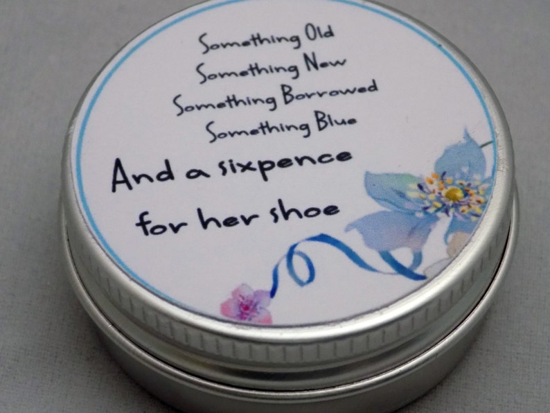 Puede incluir: Una lata de plata con una etiqueta blanca que dice "Something Old, Something New, Something Borrowed, Something Blue, And a sixpence for her shoe". La etiqueta tambi&eacute;n tiene una flor azul con un centro amarillo.
