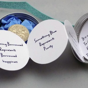 Puede incluir: Una tarjeta blanca con el texto "Something Borrowed Represents Borrowed happiness" est&aacute; unida a una lata de plata con una tapa azul. Dentro de la lata hay una moneda de plata con el texto "DEF 1955". Otra tarjeta blanca con el texto "Something Blue Represents Purity" est&aacute; unida a la lata. Una tercera tarjeta blanca con el texto "Es habitual que una novia se meta una moneda de seis peniques en el zapato izquierdo durante la ceremonia de boda" est&aacute; unida a la lata.