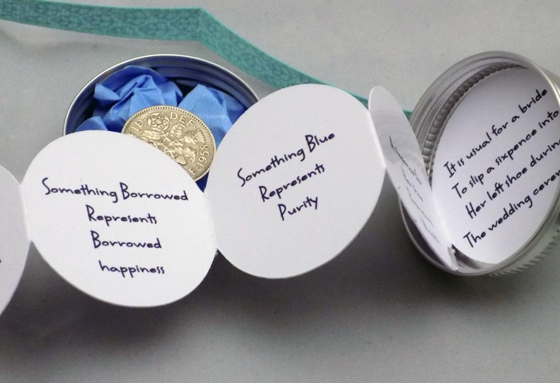 Puede incluir: Una tarjeta blanca con el texto "Something Borrowed Represents Borrowed happiness" est&aacute; unida a una lata de plata con una tapa azul. Dentro de la lata hay una moneda de plata con el texto "DEF 1955". Otra tarjeta blanca con el texto "Something Blue Represents Purity" est&aacute; unida a la lata. Una tercera tarjeta blanca con el texto "Es habitual que una novia se meta una moneda de seis peniques en el zapato izquierdo durante la ceremonia de boda" est&aacute; unida a la lata.
