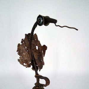 May include: A whimsical sculpture made from repurposed metal and found objects. The sculpture features a rusty metal bird with a single eye made from a bottle cap, perched on a brass bell base.
