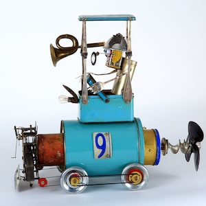 Peut inclure: Un train jouet bleu et rouge avec une hélice, une ampoule et une figurine dans la cabine. Le train porte le numéro "9" sur le côté.