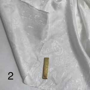 May include: White fabric with a paisley pattern. The fabric is smooth and has a subtle sheen. A ruler is placed on the fabric for scale.
