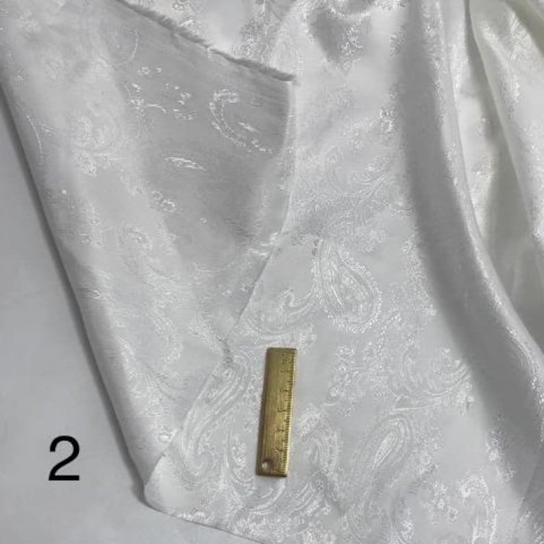 May include: White fabric with a paisley pattern. The fabric is smooth and has a subtle sheen. A ruler is placed on the fabric for scale.