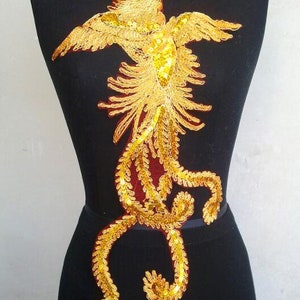May include: Gold sequined phoenix applique with a red velvet backing. The phoenix is in a standing position with its wings spread out. The tail feathers are long and flowing.