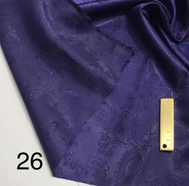 May include: A close-up of a purple fabric with a paisley pattern. The fabric is folded and has a gold measuring tape on top of it. The number 26 is visible in the bottom left corner of the image.