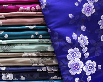 May include: A stack of fabric swatches in various colors and patterns, including teal, blue, green, pink, and purple. The fabric has a floral print with white flowers and dark green leaves. The fabric is folded neatly and stacked on top of each other.