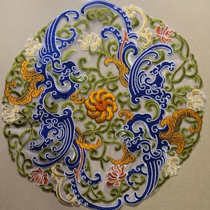 Puede incluir: Una tela redonda, blanca, bordada con un diseño floral azul, verde, amarillo y naranja. El diseño presenta patrones en espiral y flores estilizadas.