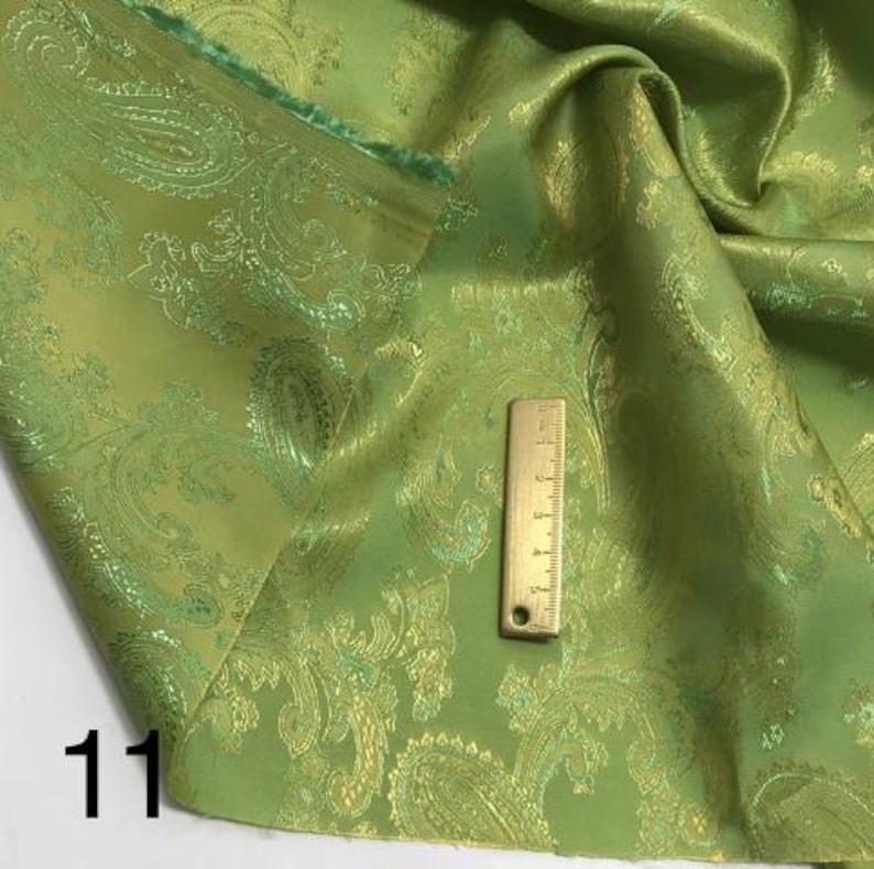 May include: Green paisley fabric with a gold metallic sheen. The fabric is folded and a ruler is placed on top of it. The number "11" is written in the bottom left corner of the image.