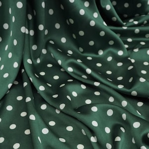 May include: Close-up of a dark green satin fabric with a pattern of white polka dots. The fabric is draped, creating folds and shadows that highlight the sheen of the material. The polka dots are evenly spaced.