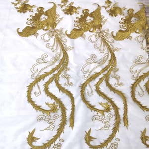 May include: Gold embroidered phoenix design on white fabric. The embroidery features detailed phoenix figures with flowing tail feathers and floral accents. The design is likely for decorative purposes, such as embellishing clothing or home decor items.