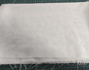 May include: White fabric with a subtle embossed floral pattern. The fabric is folded and has a frayed edge.