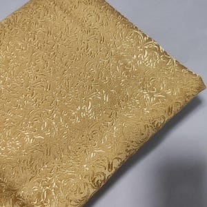 Op de afbeelding: Een gouden stof met een reliëf gegraveerd bloemenpatroon. De stof heeft een zachte, fluweelachtige textuur.