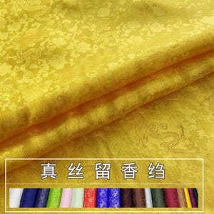 May include: Close-up of a folded, golden-yellow silk fabric with a subtle floral and dragon pattern. The fabric has a luxurious sheen and texture. The image includes a rectangular box with Chinese characters and a row of colorful fabric samples.