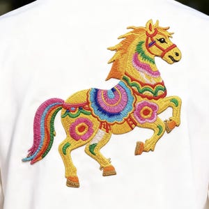 May include: White shirt featuring a colorful embroidered horse design on the back. The horse is yellow with a rainbow mane and tail, and is adorned with floral and geometric patterns in various colors.