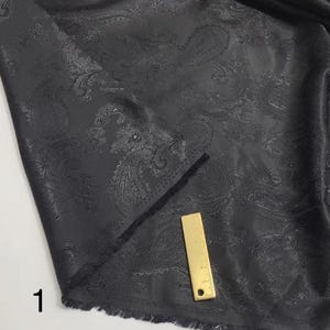 May include: Black paisley patterned fabric with a subtle sheen. The fabric has a fringed edge and a ruler is placed on top of it.