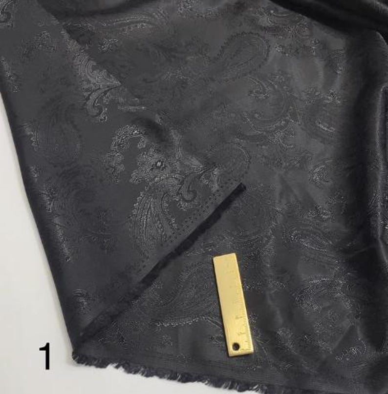 May include: Black paisley patterned fabric with a subtle sheen. The fabric has a fringed edge and a ruler is placed on top of it.