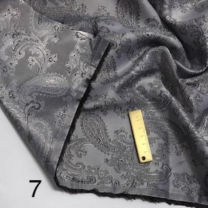 May include: Gray paisley patterned fabric with a silver sheen. The fabric is folded and a ruler is visible in the image.