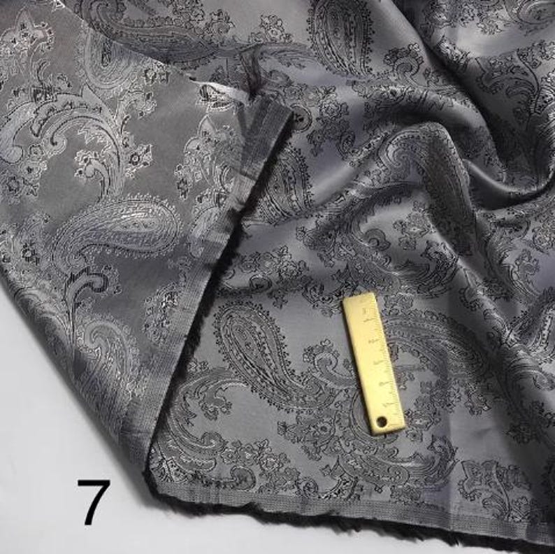 May include: Gray paisley patterned fabric with a silver sheen. The fabric is folded and a ruler is visible in the image.