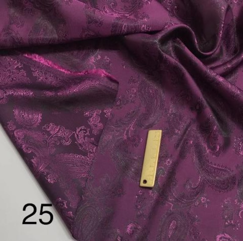 May include: Purple paisley patterned fabric with a black paisley design. The fabric is folded and a ruler is visible in the image. The number 25 is visible in the bottom left corner of the image.