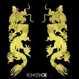 Puede incluir: Dos apliques dorados de dragones con detalles intrincados, sobre un fondo negro. Cada dragón está en una pose sinuosa, con escamas detalladas y rasgos fluidos. Dimensiones: 83 x 29 cm.