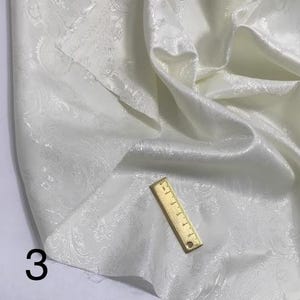 May include: White fabric with a paisley pattern. The fabric is smooth and has a subtle sheen. A ruler is lying on top of the fabric.