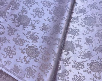 May include: Close-up of a light purple fabric with a repeating pattern of stylized floral and geometric designs in a silver-gray color. The fabric is folded, showing the texture and sheen.