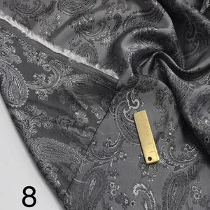 May include: Gray paisley patterned fabric with a silver sheen. The fabric is folded and draped, showing the texture and pattern. The number "8" is visible in the lower left corner of the image.