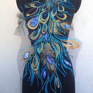 Puede incluir: Un diseño de pluma de pavo real azul y dorado bordado sobre una tela transparente. El diseño presenta detalles intrincados y un estilo fluido y elegante.