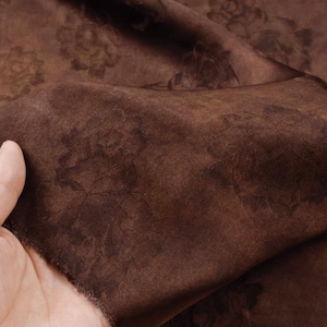 May include: A close-up of a piece of brown fabric with a floral pattern. The fabric is a rich, dark brown color with a subtle sheen. The floral pattern is a darker shade of brown, creating a tone-on-tone effect. The fabric is soft and appears to be draped.