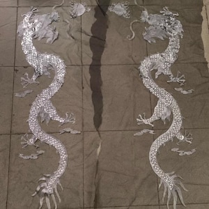 Könnte beinhalten: Zwei silberne Pailletten-Drachen-Applikationen auf einem schwarzen Netzstoff. Die Drachen sind einander zugewandt und haben ihre Schwänze eingerollt.