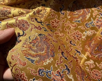 Könnte beinhalten: Goldfarbener Stoff mit einem detaillierten Stickmuster aus Drachen und Blumen. Der Stoff hat einen dezenten Glanz und fühlt sich luxuriös an.