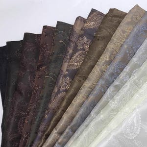 May include: A collection of fabric swatches in various shades of brown, grey, and white with paisley patterns. The fabrics are arranged in a fan shape, with the lightest fabric at the bottom and the darkest fabric at the top.