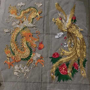 May include: Two embroidered patches featuring mythical creatures. One patch depicts a golden dragon with orange and green accents, surrounded by clouds. The other patch shows a golden phoenix with red flowers and green leaves.
