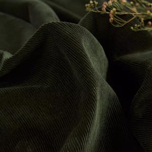 May include: A close-up of dark green corduroy fabric with a soft, textured surface. The fabric is folded and draped, creating a sense of depth and dimension.