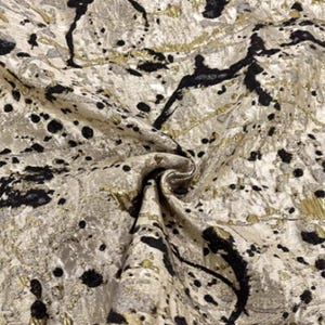 May include: Close-up of a textured fabric with a pattern of black splatters and gold accents on a cream background. The fabric is folded, creating shadows and highlighting the texture. The overall effect is one of luxury and artistic design.