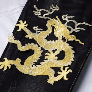 May include: Close-up of black denim fabric featuring a large, gold embroidered dragon design. The dragon is intricately detailed with a textured appearance, showcasing its scales and claws. The embroidery has a metallic sheen.