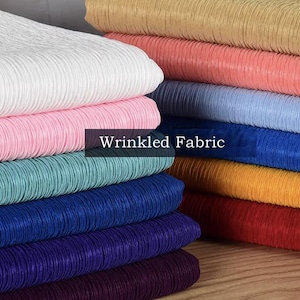 Op de afbeelding: Een stapel kleurrijke, gekreukelde stof in verschillende tinten, waaronder wit, roze, blauwgroen, blauw en rood. De tekst "Wrinkled Fabric" wordt op een donkere achtergrond weergegeven. De stof heeft een geribbelde textuur.