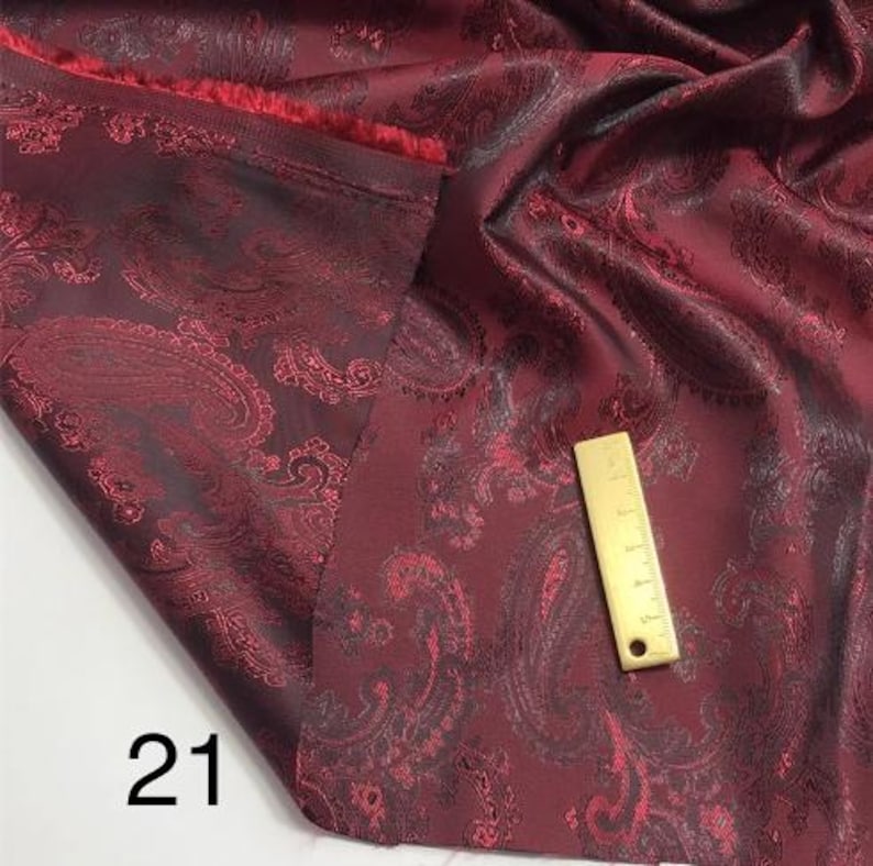 May include: Burgundy paisley fabric with a gold-coloured ruler on top. The fabric has a subtle sheen and a textured pattern.