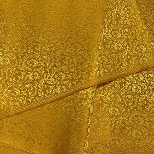 May include: A close-up of a gold fabric with a repeating floral pattern. The fabric is folded in half, showing the pattern on both sides.