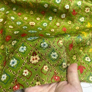 Puede incluir: Primer plano de una tela verde con un patrón repetido de formas florales y geométricas. El diseño presenta pequeñas flores en blanco, rojo y azul, dispuestas dentro de formas de diamante y cuadrifolio. La tela tiene un aspecto texturizado.