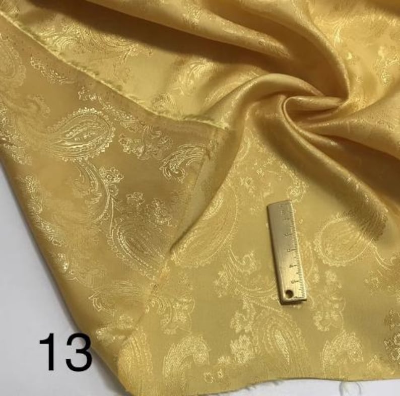 May include: A close-up of a gold paisley patterned fabric. The fabric has a satin-like sheen and is folded to show the pattern.