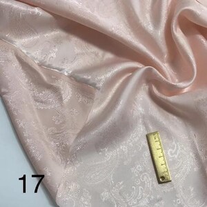 May include: A pale pink fabric with a white paisley pattern. The fabric is smooth and silky. A ruler is included for scale.